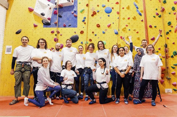 Group photo of people in climbing gear standing in front of an indoor climbing wall, wearing Settle t-shirts